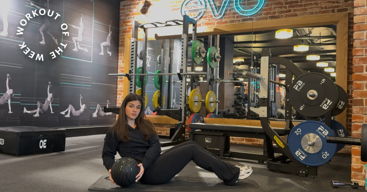 Entraînement de la semaine : entraînement de force du tronc de 15 minutes - Femme assise sur un tapis tenant un medecine ball dans une salle avec rack à squat et poids derrière